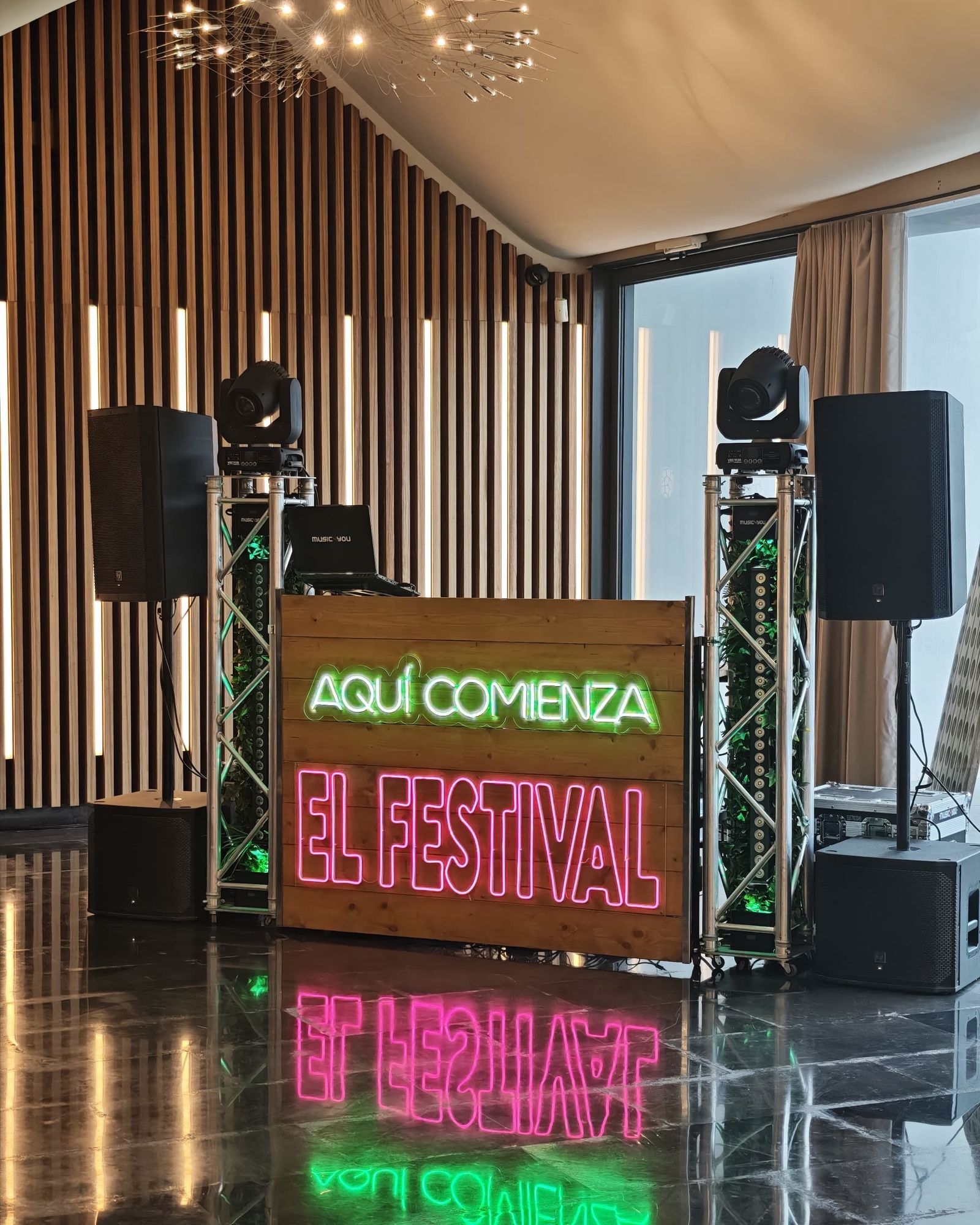 Cabina de DJ de Music4you con el rótulo de neón «Aquí comienza el festival», dos tótems con cabezas móviles y altavoces en un salón